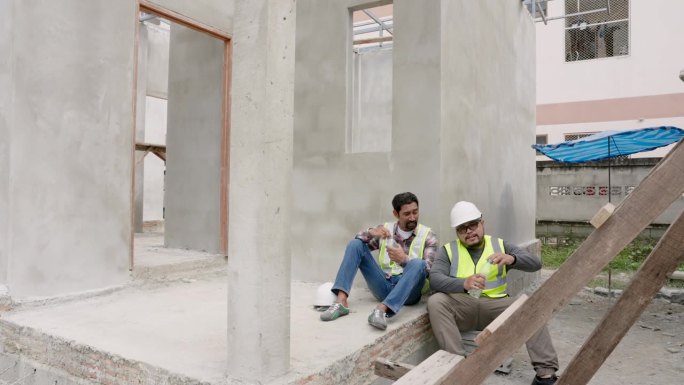 印度工头和建筑师坐下来休息。身穿绿色反光背心的男子摘下头盔。在工地上干活后，靠在水泥墙上坐着，喝点水