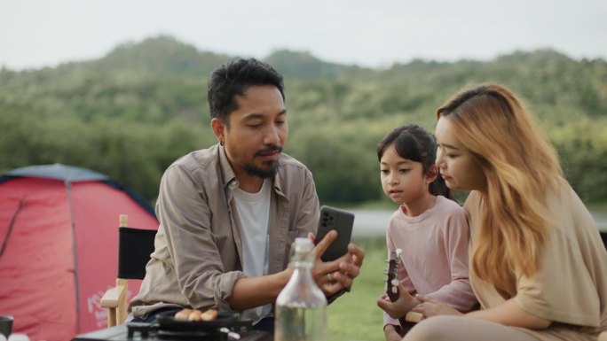 父亲在智能手机上播放有关音乐节的视频。妈妈和孩子们兴致勃勃地坐在一起露营。