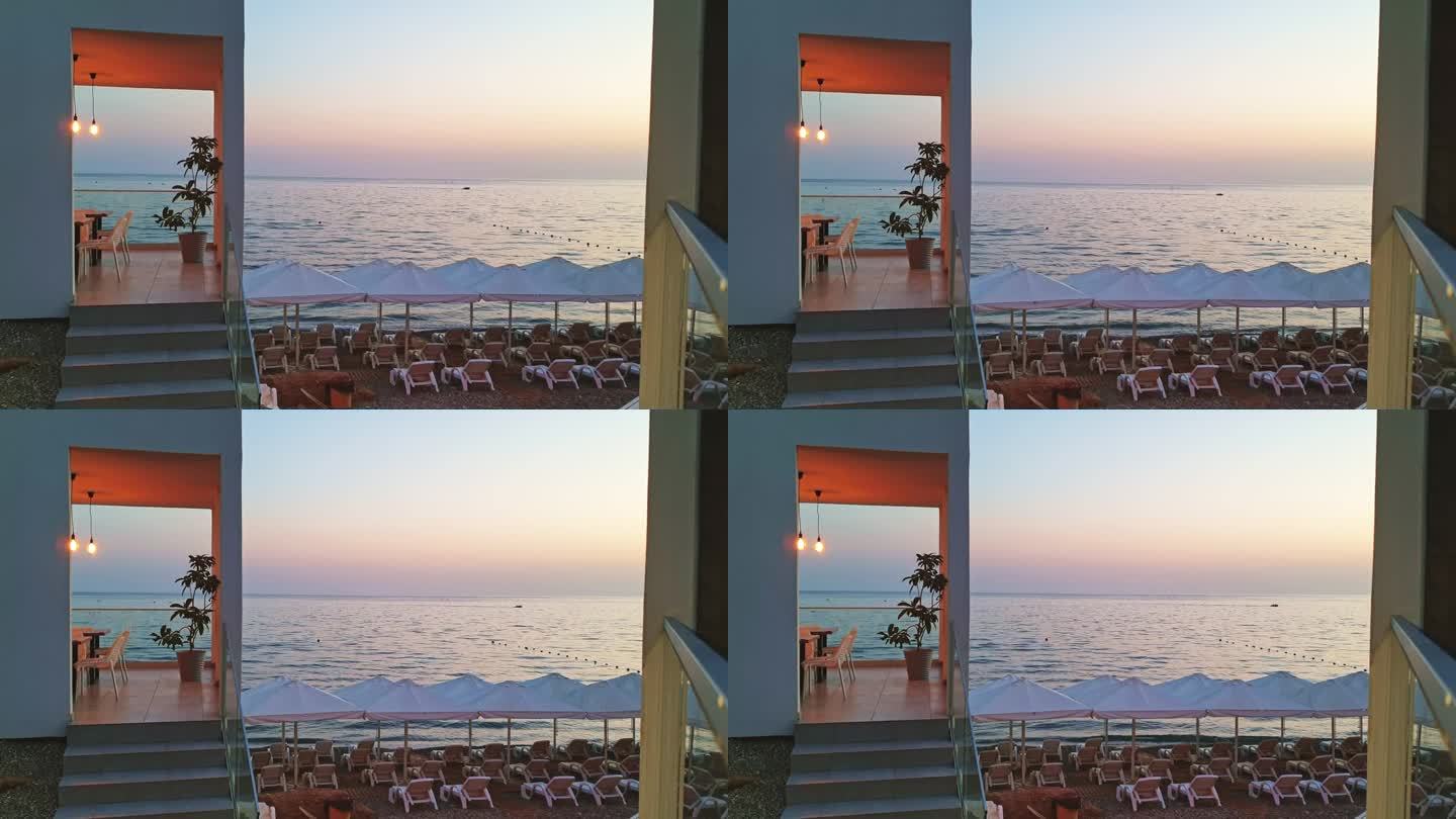 日落时的海边度假胜地。粉红色的晚霞倒映在大海上。沙滩上，白色的遮阳伞和躺椅，旁边的白色咖啡馆有无花果