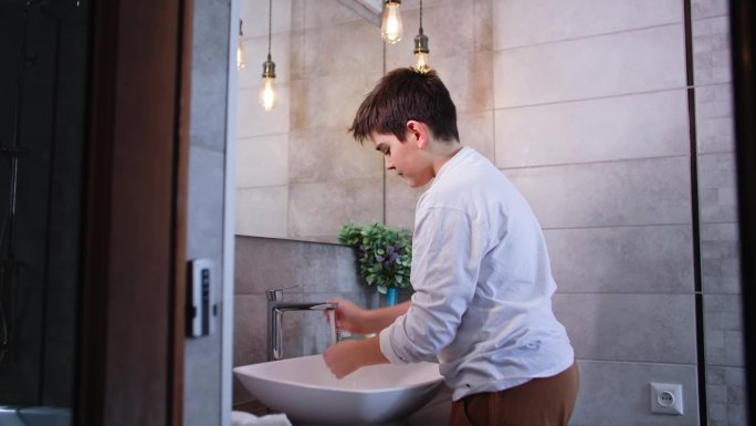 个人卫生，可爱的少年站在浴室水槽旁，用肥皂洗手，用水冲洗掉泡沫