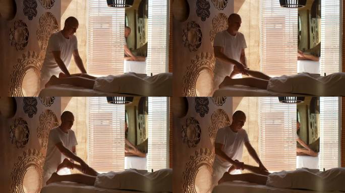 专业男按摩师，双手有力，对躺在窗边按摩床上的运动员小腿肌肉进行运动按摩。