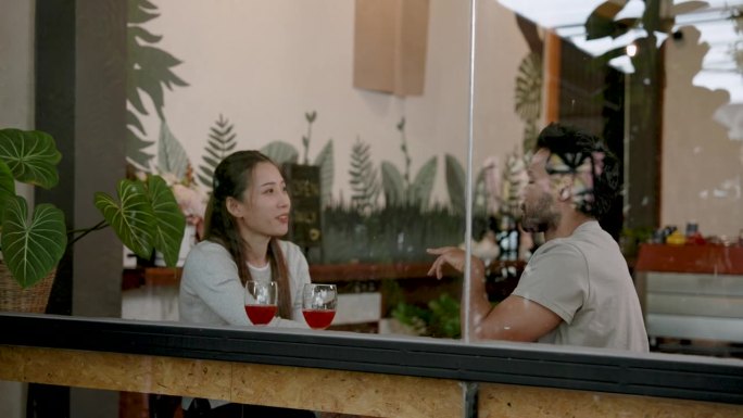 透过玻璃看到一个美丽的亚洲女人和一个印度男人。坐在咖啡店的窗边喝葡萄酒。在一天的空闲时间愉快地聊天。
