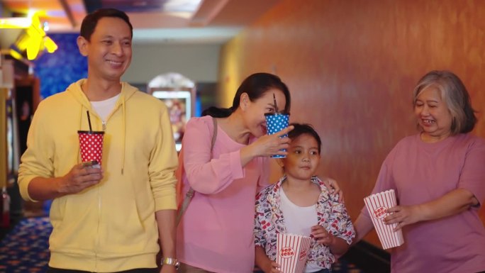 快乐的亚洲家庭带着爆米花和饮料准备一起去电影院看电影。亚洲家庭拿着碳酸饮料和一桶爆米花去电影院看电影