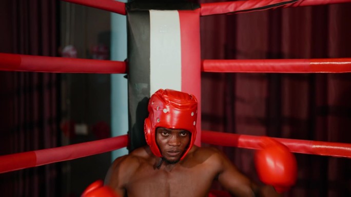 非洲年轻的成年拳击手，带着一套拳套，疲惫地坐在拳击台的转盘上。