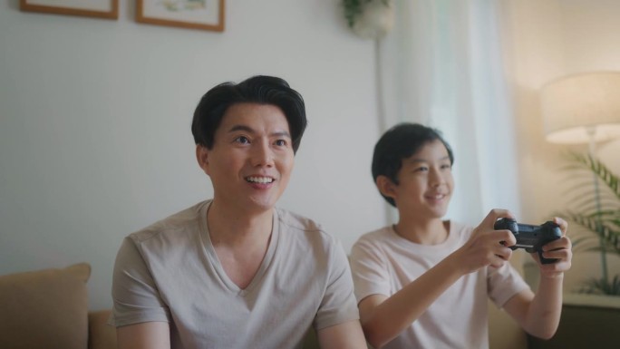 亚洲父亲和孩子在家里用摇杆玩电子游戏，在度假时一起聊天。