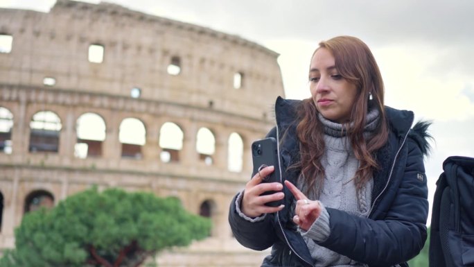 在意大利罗马的历史建筑斗兽场前，带着行李和保暖衣服的年轻拉丁女游客在多云的天空中玩着智能手机
