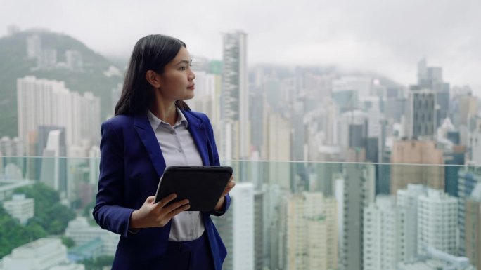 年轻的女商人独自站在外面，用着平板电脑，背景是香港岛的建筑。