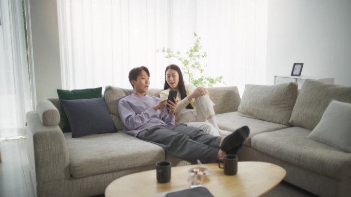不同的韩国夫妇在明亮舒适的客厅沙发上玩智能手机，一起享受时光。寻找度假目的地的青年男女
