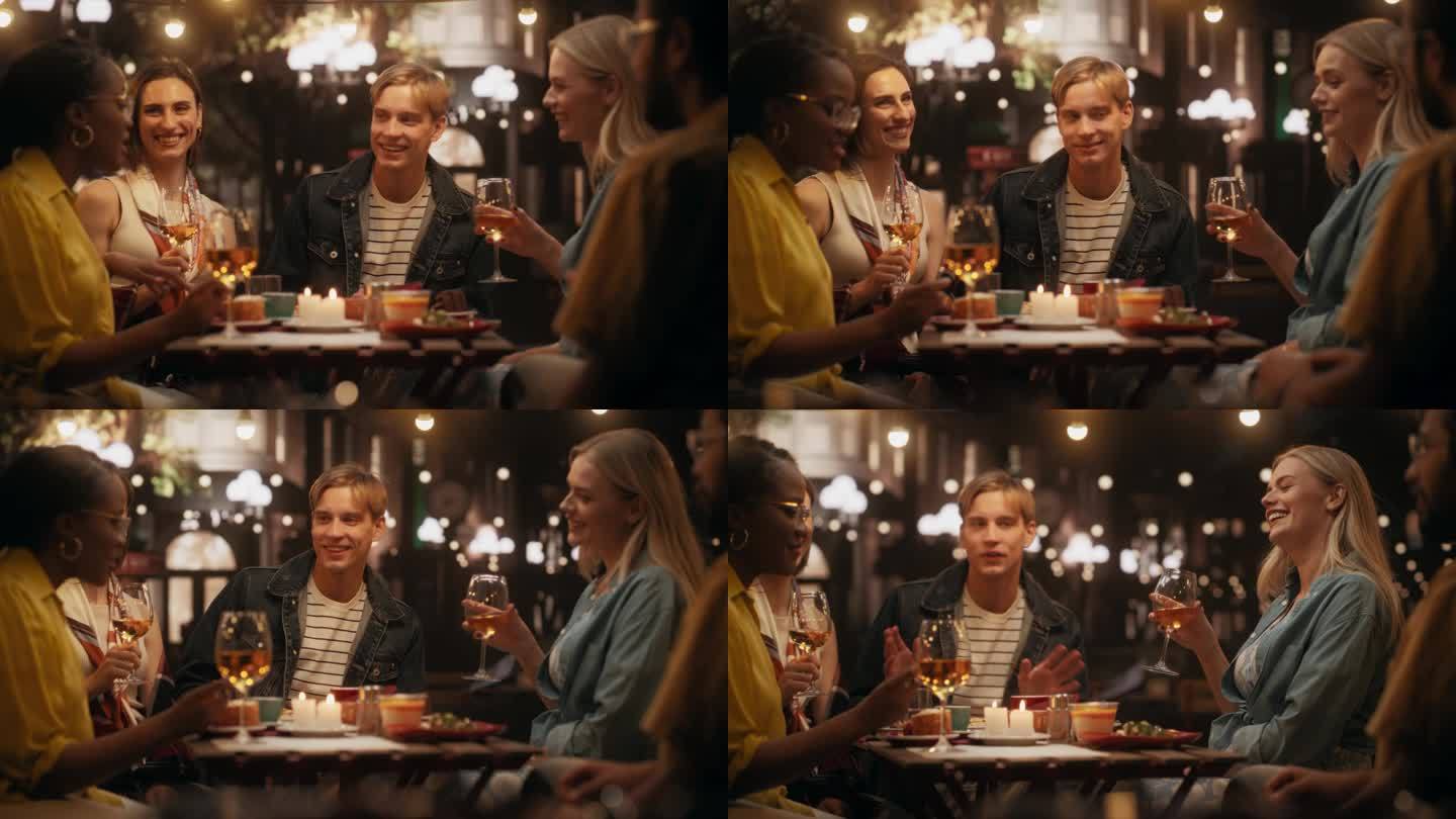 一群不同的朋友在街边咖啡馆享受夜生活。年轻的女人和男人坐在桌子后面，有乐趣和快乐的谈话。白人男子分享