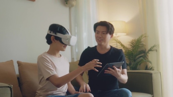 一个亚洲家庭，爸爸使用笔记本电脑，儿子戴着虚拟现实眼镜，头戴式耳机，坐在客厅开始玩电子游戏。