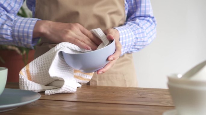 女性用白色的华夫饼织毛巾擦盘子。在一个质朴的厨房里，穿着米色围裙的女人在擦拭洗过的盘子