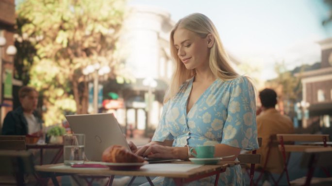 美丽的女人在夏日舒适的咖啡馆露台上使用笔记本电脑。金发碧眼的年轻女子在一个蓝色的连衣裙与花卉图案喝咖