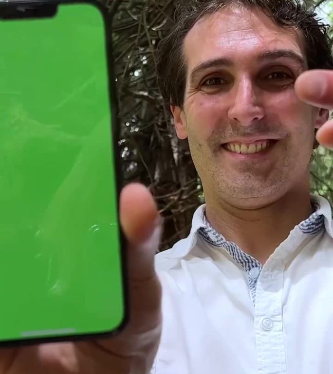 一个男人手里拿着绿屏色度键的手机，他穿着白衬衫站在森林里向镜头展示手机
