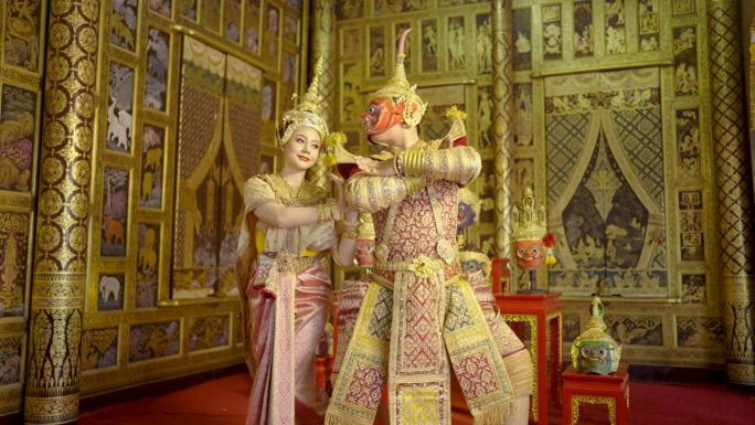 泰国经典的蒙面男子从拉玛金角色带着红色面具与美丽的亚洲女人跳舞，穿着泰国传统服装，呆在公共场所墙上的