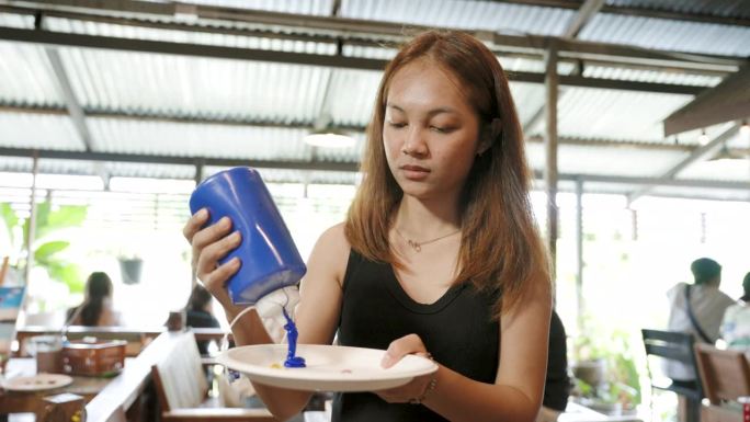 画像中年轻女子手里拿着水彩瓶，正用手把蓝色的瓶子挤进纸盘，把原色挤进纸盘，如果要用其他颜色，就用原色