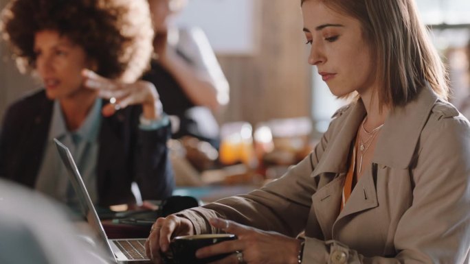 年轻女子使用笔记本电脑在咖啡馆工作，在智能手机社交媒体上分享信息，享受坐在繁忙的餐厅喝咖啡