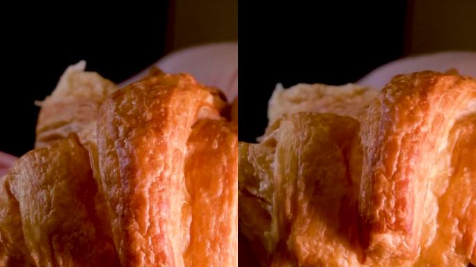 女性双手掰开新鲜温暖的牛角面包美味的早餐黑色背景上的自制蛋糕碎屑落在石头桌上是可见光结构多孔糕点面包