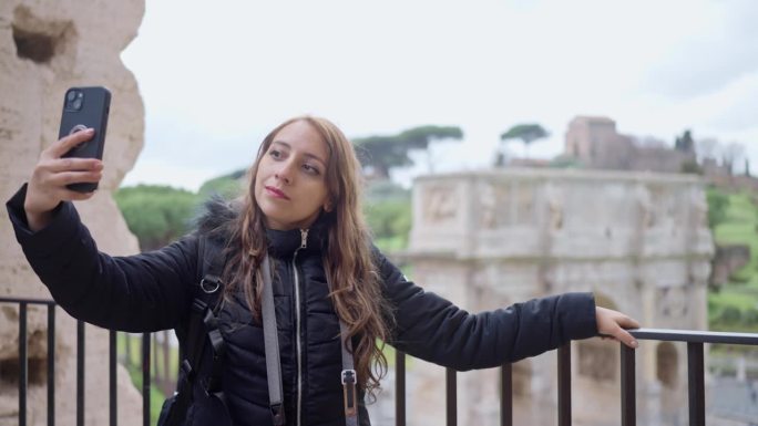 年轻的拉丁女性游客穿着保暖的衣服，拿着专业相机，在意大利罗马斗兽场用智能手机自拍，一边欣赏历史建筑，