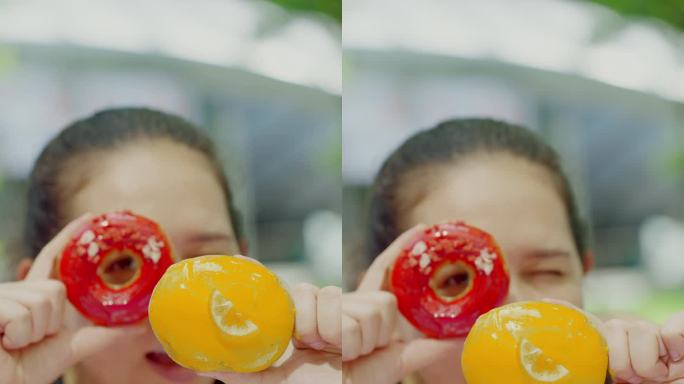 特写:看着镜头:亚洲中年妇女双手拿着两个红色和黄色的甜甜圈，一边玩一边移动甜甜圈，一边开心地跳舞。她