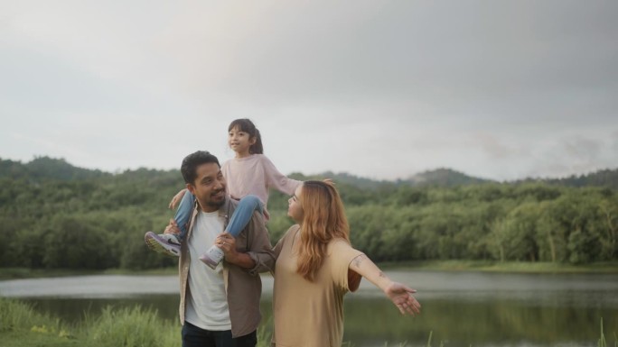 幸福的一家人，女儿在爸爸的肩膀上走在草地上，妈妈和女儿像小鸟一样在空中张开双臂。