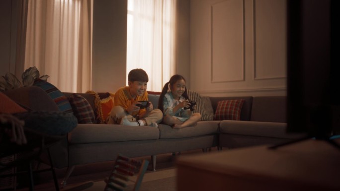 可爱的小兄弟姐妹在舒适的客厅沙发上的游戏机上玩电子游戏。韩国兄弟姐妹玩游戏，一起赢，击掌。孩子们放学