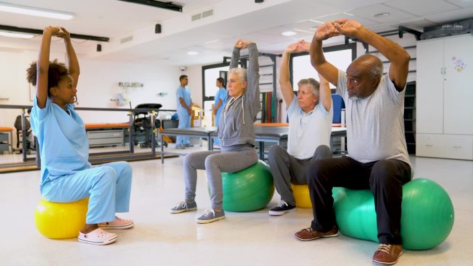 一群老年人坐在健身球上，跟随一位黑人女理疗师给他们做的练习