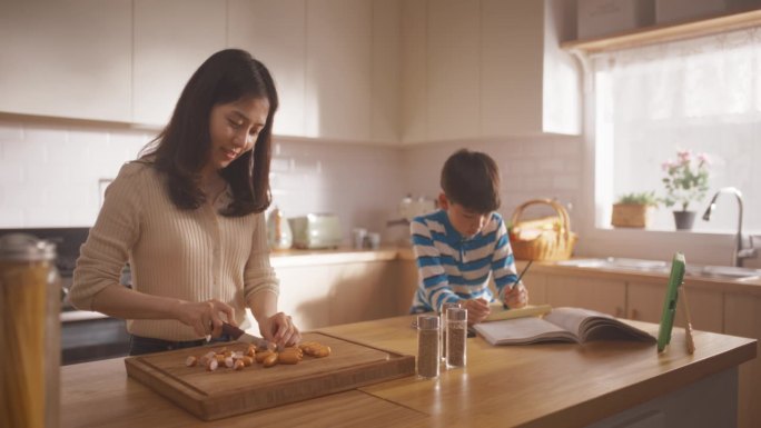 一个韩国女人和她的儿子在家里的厨房里的肖像:美丽的母亲准备做午餐，同时和她旁边做作业的可爱的孩子说话