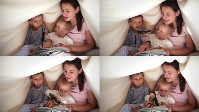 幸福的一家人穿着睡衣，坐在毯子下面，拿着手电筒看书。家人在一起，养育子女，快乐的童年和娱乐