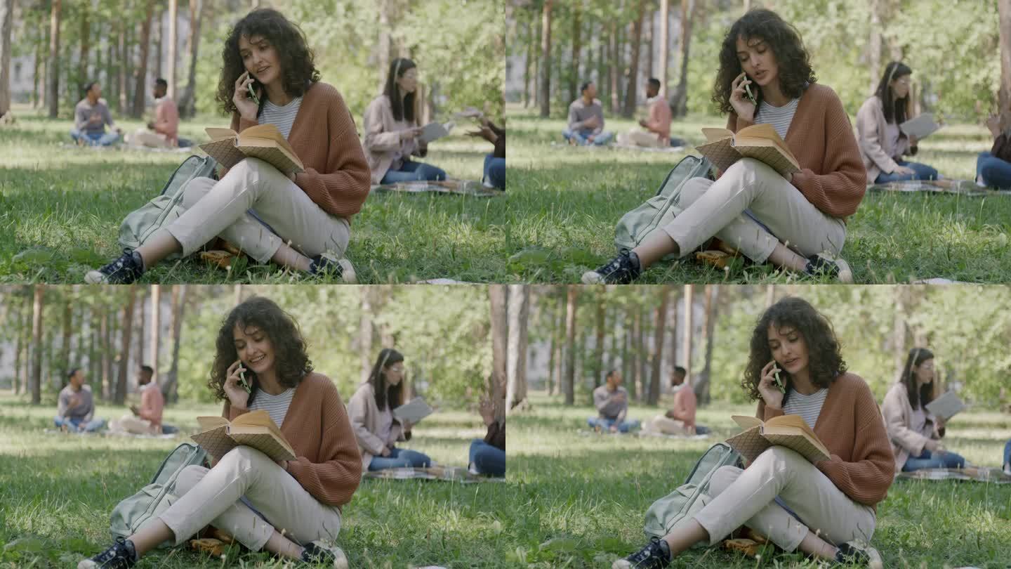 快乐的女学生坐在公园的草地上看书和打电话的画像