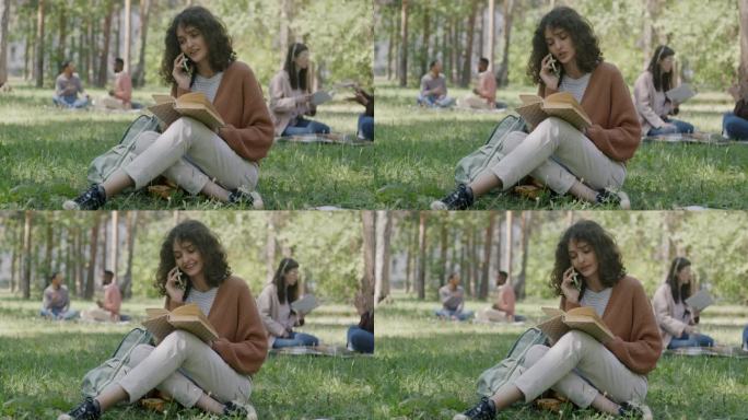 快乐的女学生坐在公园的草地上看书和打电话的画像