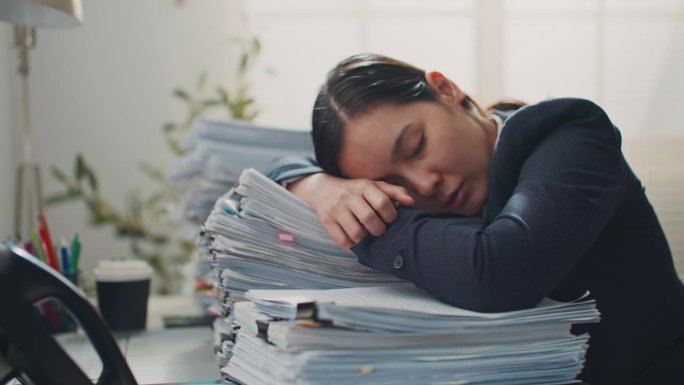 疲倦困倦的年轻女商人在办公桌上堆着一叠文件睡着了