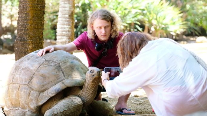 在毛里求斯岛的动物园自然公园里，一位游客妈妈用手机给儿子和世界上最大的陆龟之一——阿尔达布拉巨型陆龟