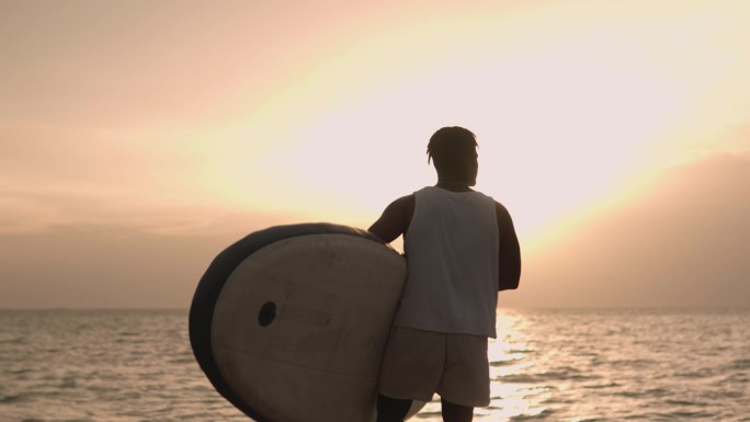 一个非裔美国少年手持冲浪板在海滩上走向大海。