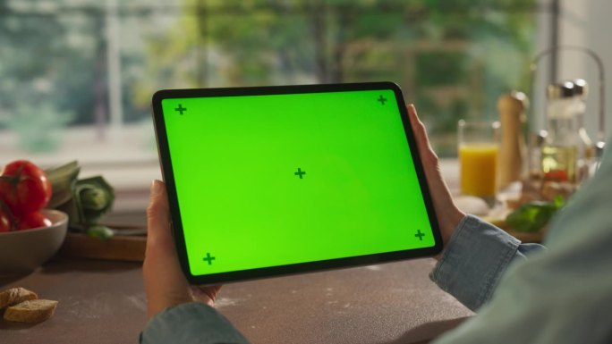 一名匿名女子手持平板电脑，背景是厨房的桌子。数字设备与绿色屏幕色度键模板的数字营销，在线服务，应用程