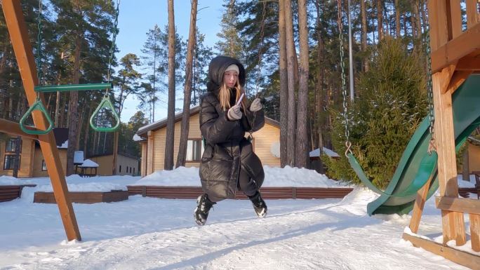 女孩穿着羽绒服，戴着帽子，在下雪的公园里用智能手机荡秋千。