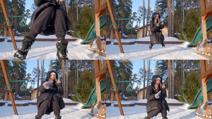女孩穿着羽绒服，戴着帽子，在下雪的公园里用智能手机荡秋千。