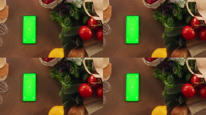 智能手机的顶视图与模拟绿色屏幕显示。一个设备与蔬菜垂直躺在厨房桌子上的静态镜头。在线数字营销和内容创