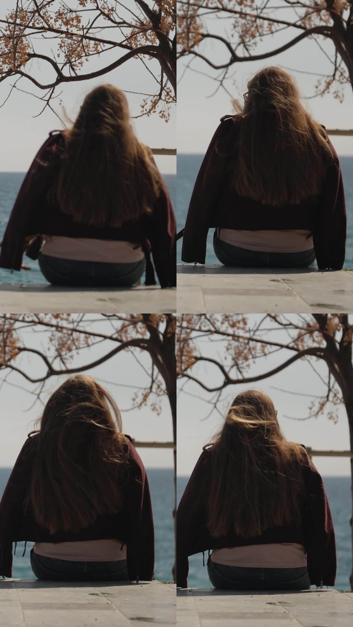 一位年轻女子坐在海边的混凝土上，望着远方。风吹乱了她的长发。垂直视频。