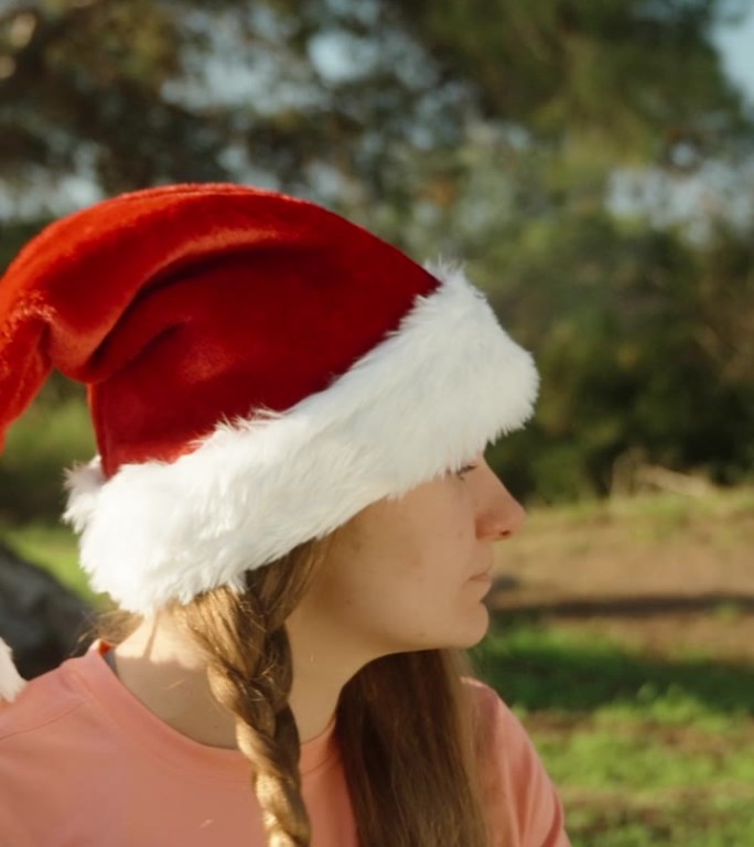 我把头发编成一个发髻，一个戴着红色圣诞老人帽的年轻女人。圣诞节在森林里做瑜伽。垂直视频。