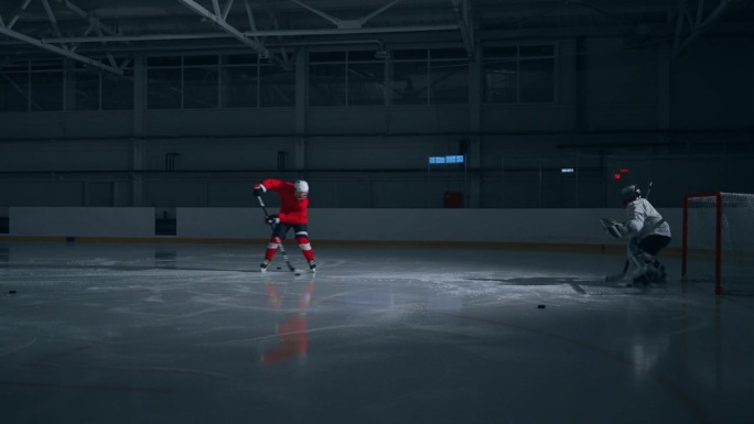 一名穿着红色运动衫的熟练曲棍球运动员在黑暗的冰场上训练，表现出快速的动作，并向守门员进球