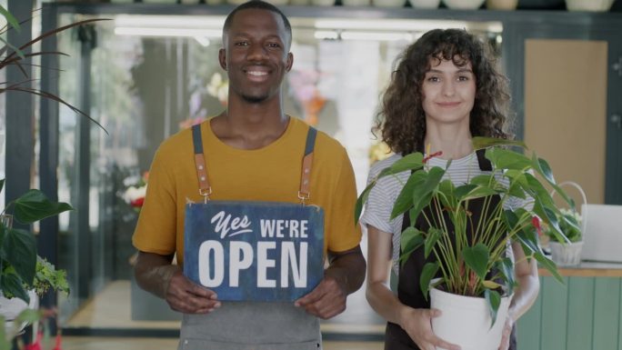 非裔美国男人和白人女人的肖像，拿着是的，我们是开放的板和盆栽植物站在花店微笑