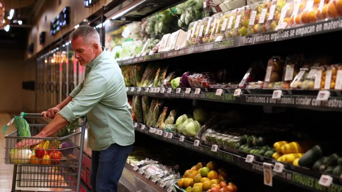 一个成熟的男人推着一辆手推车，旁边放着冷冻的有机蔬菜，抓了一些放到购物车里