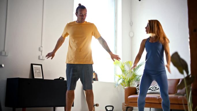 SLO MO Fit年轻夫妇在家锻炼时做深蹲跳，一起在客厅锻炼。他们停顿了一下，笑了笑才继续说下去。