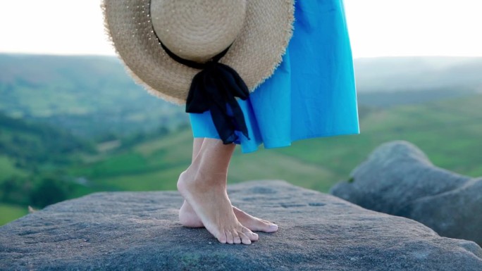 一名亚洲女子身穿蓝色连衣裙，戴着一顶帽子，手里拿着薰衣草花束，站在夏日日落的山顶上