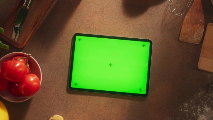 一个平板电脑与模拟绿色屏幕显示的顶部向下缩放视图。数字设备模板躺在木制的厨房桌子上，周围是健康的食物