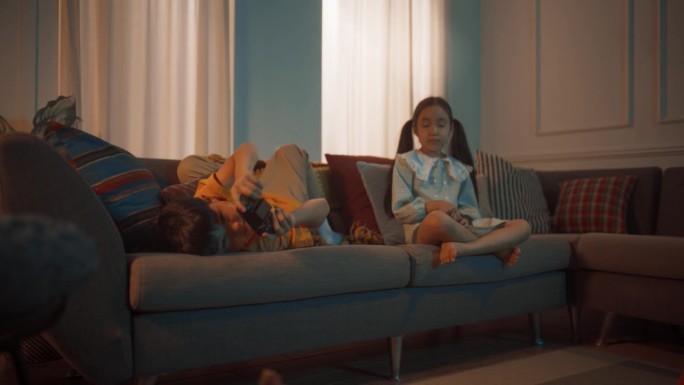 可爱的小兄弟姐妹在舒适的客厅沙发上的游戏机上玩电子游戏。韩国兄妹在晚上一起玩游戏。孩子们放学后在家玩