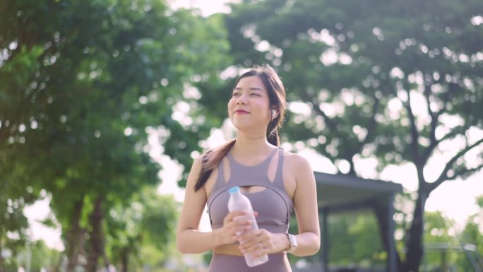 亚洲运动年轻成年女性慢跑者早上喝水，年轻女性从瓶子里喝水。女性在锻炼或运动后喝水。