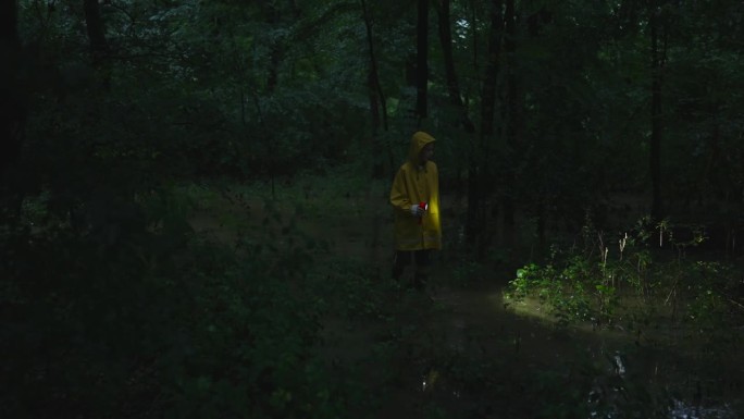 穿着雨衣，带着手电筒的男孩在雨季的森林里走在洪水泛滥的平原上