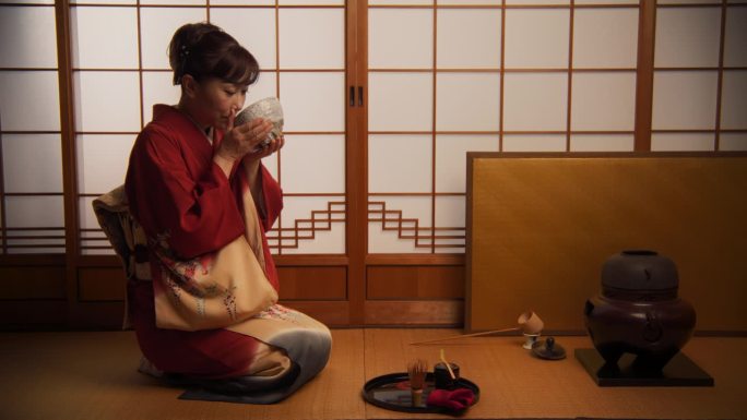 日本成年女性坐在榻榻米地板上准备抹茶绿茶。身穿传统红色和服、饰有花卉的亚洲妇女正在用碗品茶