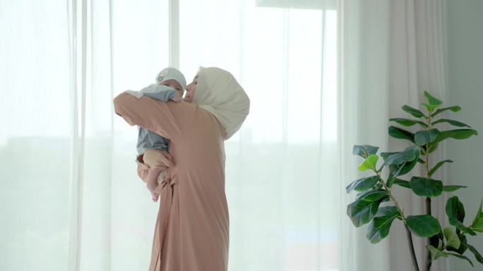 一名穆斯林妇女抱着两个月大的孩子在卧室里微笑，她把孩子放在肩上，戴着头巾，目的是让孩子更舒适、更快乐